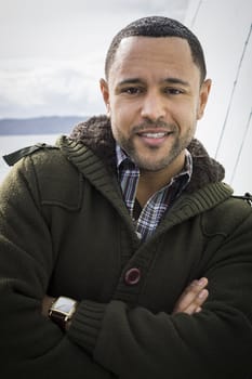 Portrait of young black man with smile and arms crossed standing on boat deck, leaning against wall with water in the background.