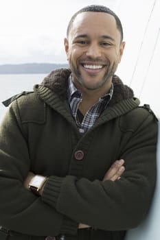 Young black man with smile on boat deck, leaning against wall with water in the background.