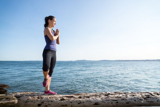 Young women on beach in upright yoga pose with water in background. She is posing in the Tree pose.