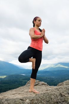 Young woman in yoga pose on top of mountain with beautiful vista in background.