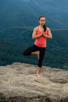 Young woman in yoga pose on top of mountain with beautiful vista in background.