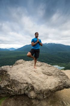 Young man in yoga pose on top of mountain with beautiful vista in background.