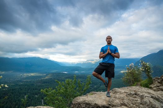 Young man in yoga pose on top of mountain with beautiful vista in background.