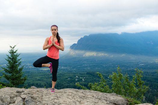 Young woman in yoga pose on top of mountain with beautiful vista in background.