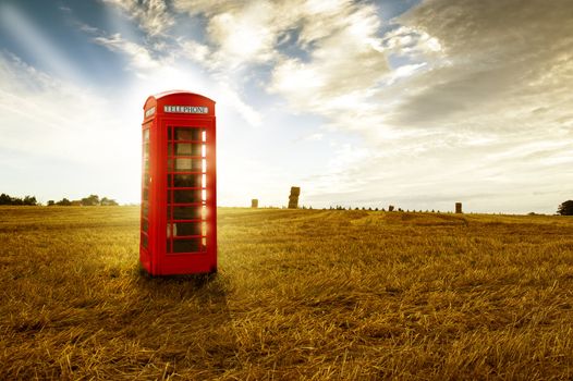 Old-fashioned traditional red telephone booth or public payphone standing in an open deserted field in evening light