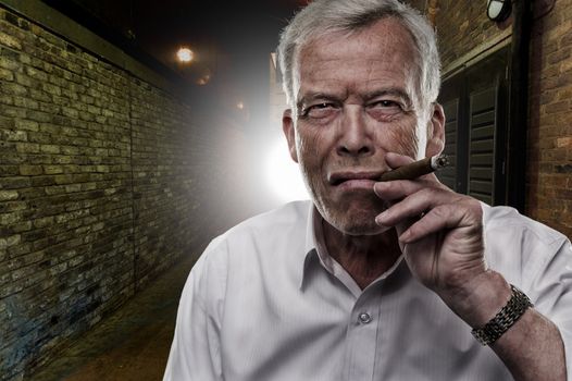 Handsome determined senior man smoking a cigar outdoors in the darkness backlit by a light on a brick building