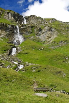 Beautiful alpine waterfall going down directly from the peaks through green  meadows