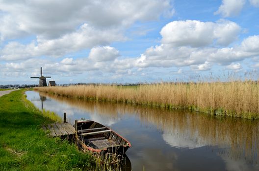 Typical landscape in Holland with an old boat in a ditch with a windmill, reeds and clouds.