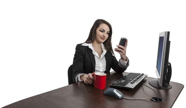 Image of a young brunette woman with a red cup of coffee checking her mobile in front of the computer at her workplace , isolated against a white background.