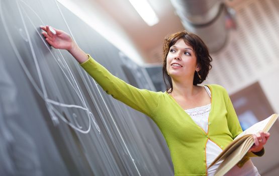 Pretty young elementary school/college teacher writing on the chalkboard/blackboard during a math class (color toned image; shallow DOF)