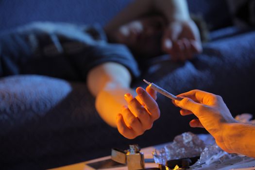 Close up of a young man who gives a joint to his friend lying on the couch