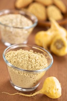 Maca powder (flour) in glass bowl with maca roots or Peruvian ginseng (lat. Lepidium meyenii) and other Peruvian maca products (oatmeal with maca and maca cookies) in the back (Selective Focus, Focus one third into the maca powder and on the front of the maca root beside the bowl)