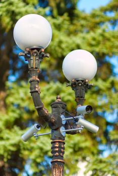 Security surveillance camera on a column with a lamp, on green tree background, selective focus