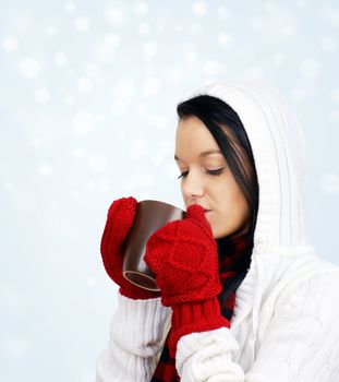 Beautiful young woman drinking comforting hot chocolate, coffee or tea in mug held with red mittens on a cold snowy winter day