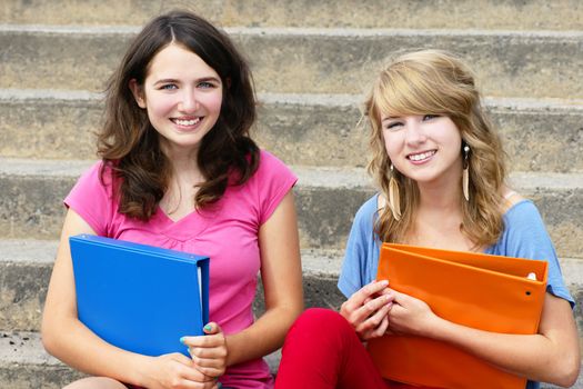 Two happy and smiling teenager girls, students or youn women, holding books while sitiing on the stairs at school.