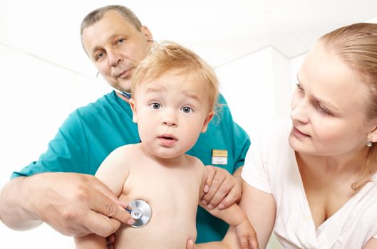 A middle-aged doctor in a green smock is listening to a baby�s back with a stethoscope in a medical study. The baby is 1�1,5 years old. He has a surprised look. The young mother is looking at her baby. Caucasian. Focus is on the boy.