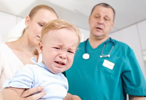 A 1,5 year-old boy is frightened and crying in a medical study. The doctor and the baby’s mother are at a loss. Shallow dof. Focus is on the boy.