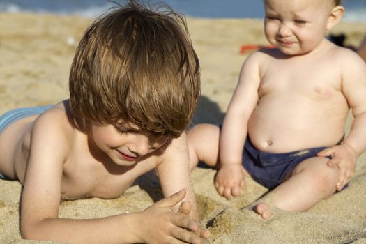 Little boy smiling and playing with sand on a beach. His baby sister looking at him. The focus is on the brother.