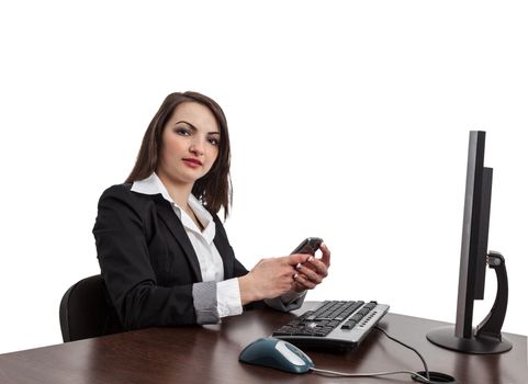 Image of a young brunette woman looking to the camera and checking her mobile in front of the computer at her workplace , isolated against a white background.