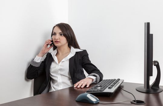 Young businesswoman at her office desk talking to a mobile phone against a white background with shadows.