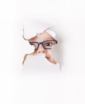 Curious kid in spectacles looks through a hole in white paper