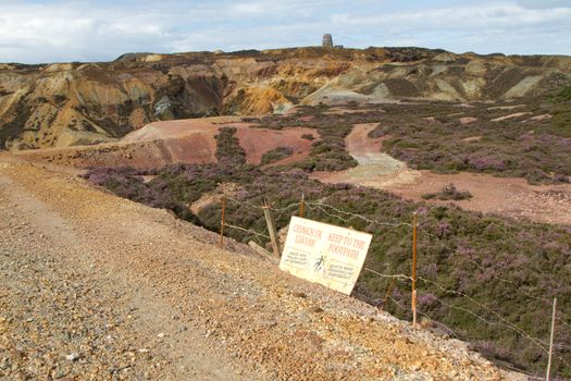 A sign with 'KEEP TO THE FOOTPATH' rests on the side of a rough track leading into an historic opencast coppermine.