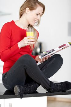 Young woman sat on kitchen work top, enjoying a glass og juice and reading a magazine