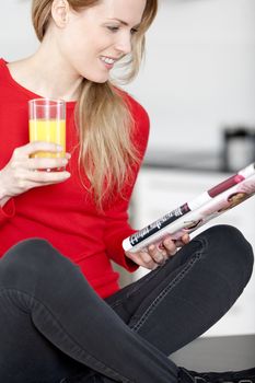 Young woman sat on kitchen work top, enjoying a glass og juice and reading a magazine