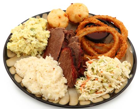 Beef brisket, grits, potato salad, cole slaw, onion rings, bbq sauce, corn muffins in a plate over white.