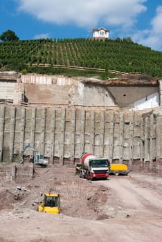 Construction site with various machinery and concrete construction into the hill and beautiful villa above vineyards with great blue sky