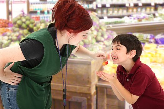 Attractive young grocery clerk sharing fresh cherries with child customer in grocery store.