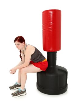 Attractive 19 year old woman sitting resting on free standing heavy bag after workout.