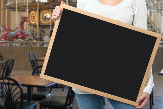 Female holding blank chalkboard in front of carousel in mall food court.