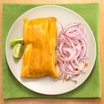 Peruvian tamale (traditionally eaten for breakfast on Sundays) made of corn and chicken and served with salsa criolla (onion salad) and limes (Selective Focus, Focus on the top of the tamale)