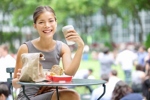 Young business woman on lunch break in City Park drinking coffee and eating sandwich. Happy smiling multiracial young businesswoman in Bryant Park, New York City, USA.