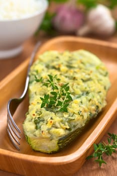 Baked zucchini stuffed with mashed potato, cheese and herbs (thyme, oregano, parsley, garlic) garnished with thyme served on wooden plate with a fork beside and a bowl of cooked rice in the back (Selective Focus, Focus on the front of the thyme garnish on the zucchini)