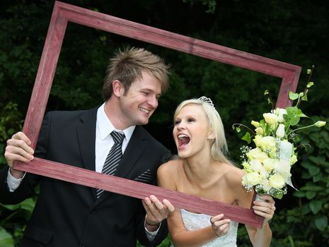 Lovely laughing wedding couple looking through a wooden picture frame