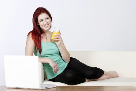 Happy young woman lying on a white sofa at home drinking Orange juice and using a laptop