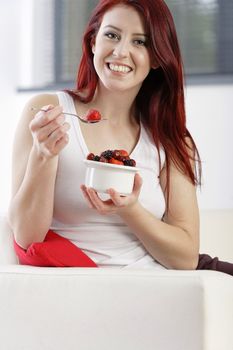 Happy young woman sat on her sofa at home enjoying a bowl of fresh fruit