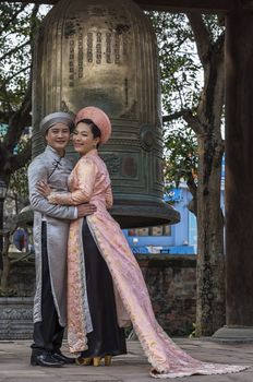 Vietnam Hanoi. Groom in grey, ping Ao Dai on bride with bronze bell background.