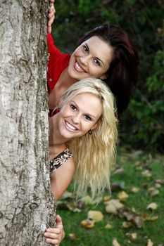 Two lovely young smiling women peeping out from behind a tree trunk