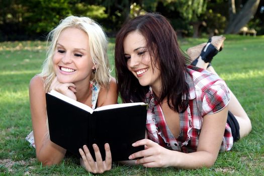 Lovely smiling brunette and blonde girls reading a book while lying on the green grass outdoors