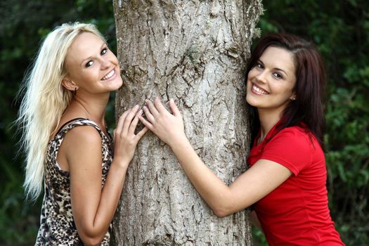 Two lovely young smiling women with arms around a tree trunk