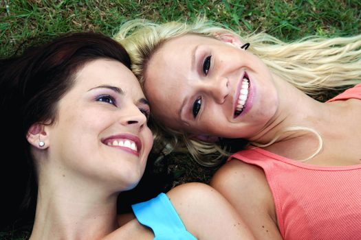 Two gorgeous smiling young women friends lying on green grass and looking up