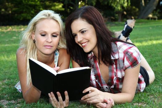 Two pretty young women reading a diary while lying on the green grass outdoors