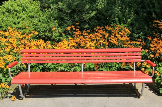 Red wooden bench in a park in front of flowers and trees