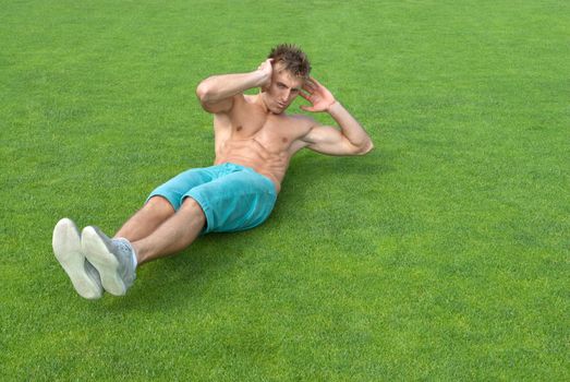Young man training outdoors and doing sit-ups on green grass.