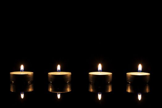 four burning tea candles on a black mirror on black background