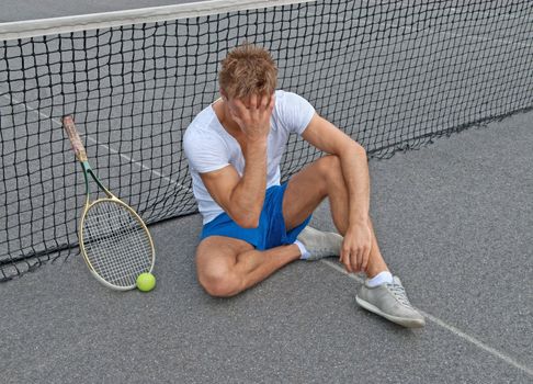 Disappointed tennis player, sitting on the ground, holding his head.