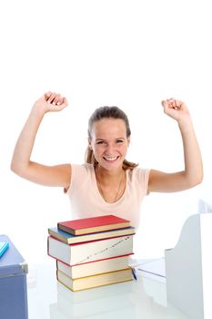 Jubilant young student sitting at her desk in front of a pile of textbooks with her hands raised in the air in celebration 